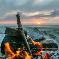 A wood fire burns on a beach with the sun near the horizon in the background.